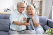 © 220 Selfmade studio - Happy senior Asian couple sitting on sofa holding smartphone smiling together with joy while looking at online content in comfortable living room