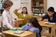 © pressmaster - Caucasian girl sitting at desk with head down being bullied by group of classmates including Caucasian boy, Asian girl, and Caucasian boy pointing and laughing in classroom