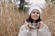 © SHOTPRIME STUDIO - Winter outdoor portrait of a smiling woman in a beige coat and white beanie among tall reeds, capturing cozy, serene fashion and natural countryside mood.