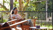 © DMegias - Mature latina woman relaxing on a porch or balcony, sitting in a wooden chair, enjoying sunlight while using a smartphone
