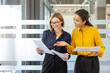 © Liubomir - Two smiling multi-ethnic businesswomen reviewing documents, discussing work, and collaborating in a bright, contemporary office environment, embodying teamwork and professional engagement
