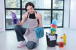 © H_Ko - In a stylish, sunlit apartment, a focused woman wears an apron and prepares to clean. She holds a spray bottle in one hand and a textured cloth in the other, surrounded by colorful cleaning supplies.