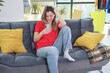 © H_Ko - A woman in a bright red shirt sits comfortably on a cozy gray couch, legs crossed, smiling as she scrolls through her phone. Yellow cushions add warmth to the space.