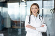© Liubomir - Young female doctor in a lab coat with stethoscope stands confidently with arms crossed, smiling warmly in a bright modern clinic, conveying professionalism and care