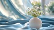 © Biancaart - White Ceramic Vase with Small White Flower Bouquet on Soft Blue Fabric in Bright Natural Light