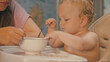© VeNN - A cute blond baby sits in a high chair, making a mess while learning to eat with a spoon. A mother watches nearby. This authentic lifestyle shot features soft lighting and a warm color palette