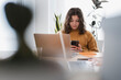 © Studio Marmellata - A young woman with curly hair is focused on her smartphone while sitting at a desk with a laptop and other work materials.