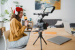 © Studio Marmellata - A young woman wearing headphones and a red hat records herself speaking into a microphone, with a camera and laptop set up for content creation.