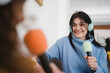 © Studio Marmellata - A young woman with headphones smiles while holding a microphone, engaging in a conversation or interview with another person.