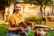 © Home-stock - Portrait of happy young woman on private farm holding chicken and smiling at camera, posing on backyard farm, promoting organic poultry farming