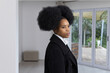 © wavebreak3 - African American woman standing in bright room near sliding doors, wearing black coat, white shirt