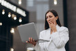 © MZaitsev - A worried businesswoman in a white suit stands outdoors near an office building, holding a laptop and touching her face while dealing with a work problem.