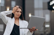 © MZaitsev - A worried businesswoman in a white suit stands outdoors near an office building, holding a laptop and touching her head while dealing with a work problem.