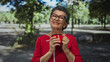 © Krakenimages.com - Woman with grey hair enjoying coffee in a park, surrounded by green trees and plants, wearing red clothing and glasses, holding a red cup with a serene expression.
