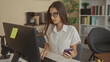 © Krakenimages.com - Woman holding smartphone and typing on keyboard in office building, wearing glasses and seated at desk with computer monitor; concentration productivity.