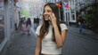 © Krakenimages.com - Woman holding smartphone to ear on street in city, smiling and talking while wearing a white crop top and jeans; casual conversation joy.