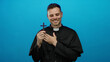 © Krakenimages.com - Priest smiling holding cross on blue background, exuding calm spirituality, dressed in black robe, hands over heart in peaceful reflection, symbolizing faith and devotion.