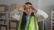 © Krakenimages.com - Young woman in a reflective vest makes a heart gesture at a volunteer center, surrounded by charity supplies, emphasizing community and kindness.