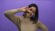 © Krakenimages.com - Young woman gestures peace sign near her eye while smiling against a vibrant purple background, showcasing playful and confident personality in a modern setting.