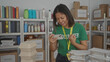 © Krakenimages.com - Young woman volunteer wearing green shirt in donation center, holding phone amid stacked containers and supplies on shelves, showcasing organized charitable efforts indoors.