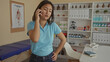 © Krakenimages.com - Woman in blue shirt talking on phone in clinic office with medical posters and shelves containing bottles and folders in background.