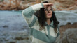 © Krakenimages.com - Young hispanic woman with curly hair and glasses gazing into the distance at a scenic beach with a rocky shoreline and clear sea.