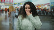 © Krakenimages.com - Hispanic woman in airport terminal indoor covering mouth with hand while wearing sweater amidst busy travelers.