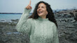 © Krakenimages.com - Woman smiling on a rocky beach with the sea in the background wearing a sweater and pointing upward, capturing a joyful outdoor moment.