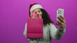 © Krakenimages.com - Woman wearing santa hat taking selfie with pink bag against vibrant pink background showcasing festive mood and joyful expression.