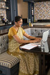 © wavebreak3 - Indian woman sitting on bench typing on laptop with notebook in modern kitchen in floral saree