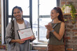 © wavebreak3 - Diverse female coworkers celebrating framed certificate at office with window plant shelf