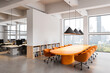 © ImageFlow - Modern office interior with orange meeting table and city view through large windows in bright open space background. 3D Rendering