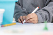 © Jeanne Sager Photo - A student's hand grips a red marker over white paper at a desk, blurred classroom behind