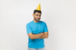 © khosrork - Portrait of satisfied handsome unshaven man wearing blue T- shirt and yellow party cone standing with crossed arms, looking at camera. Indoor studio shot isolated on gray background.