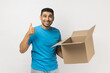 © khosrork - Portrait of smiling delighted unshaven man wearing blue T- shirt standing holding cardboard parcel, showing approved gesture, thumb up. Indoor studio shot isolated on gray background.