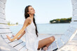 © SHOTPRIME STUDIO - Young woman sits in a white summer dress inside a circular wooden frame on a sunny beach, smiling softly as calm sea waves shimmer under a clear blue sky.
