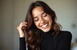 © Maryna - Attractive brunette woman laughs joyfully indoors. Her eyes are closed, head tilted, showing a broad toothy smile. She touches her wavy brown hair, feeling happy and confident in casual attire.