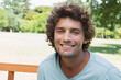 © wavebreak3 - Adult man smiling while sitting on wooden park bench in sunny park wearing light blue T-shirt