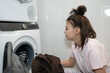 © olga_sova - A woman is sorting clothes to put into a washing machine in her home laundry area. She is focused on the task and surrounded by clean, white tiles.
