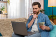 © Andrii Iemelianenko - Young man at home holding money cash dollars, looking thoughtfully at laptop screen while deciding on expenses. Caucasian guy sitting on sofa, considering purchases, investments, or financial planning