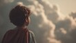 © CYBERPINK - Young boy standing in front of a cloudy sky. he is facing away from the camera, with his back towards the viewer. the boy has short, curly hair and is wearing a red cape over his shoulders.