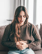© luciano - Young woman with stomach pain sitting on the couch alone at home, holding her stomach, touching and massaging her abdomen. She has an unsightly, sickly face, and feels uncomfortable and has diarrhea.
