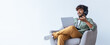 © Prostock-studio - A young man sits comfortably in a chair, wearing headphones and smiling. He uses a laptop for an online meeting. The background is simple and bright, creating a relaxed setting.