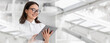 © Prostock-studio - A woman is smiling while using a tablet in a modern office environment. She is dressed in a white shirt and holds the tablet in both hands. The office has large windows.