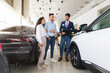 © Prostock-studio - Happy young couple buying new auto at car showroom, handsome middle eastern man sales manager assisting wealthy family, standing by nice white automobile, having convesation, full length