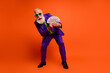 © deagreez - Eccentric elderly man in purple suit holds cash and smiles in bright orange studio for retro style promo