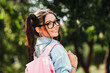 © deagreez - Young woman with glasses smiles while carrying pink backpack enjoying a sunny outdoor day in the park ready for shopping and casual stylish lifestyle