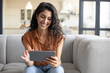 © Prostock-studio - Portrait of positive young woman using digital tablet, sitting on comfy sofa at home, free space. Cheerful millennial female browsing internet, watching video, having video chat with friend indoors