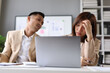© Wasan - Two Asian businesspeople expressing concern and experiencing stress while working on a laptop, analyzing financial reports in a busy office.
