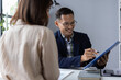 © Wasan - Asian businessman pointing at a financial report with a pen while discussing details with a colleague during a meeting in a corporate office, highlighting teamwork and strategy.